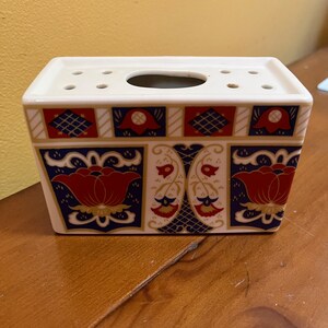 May include: Rectangular ceramic floral arrangement holder. The white holder features a decorative pattern of red, blue, and gold floral designs. The top has several small holes and a larger oval opening.