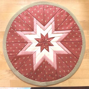 Könnte beinhalten: Eine runde, gesteppte Stoffmatte mit einem zentralen Sternmuster. Das Design besteht aus Segmenten aus altrosa Stoff mit kleinem Blumenmuster, die sich mit cremefarbenen und hellrosa Streifen abwechseln, eingefasst von einem strukturierten salbeigrünen Rand.
