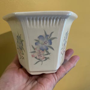 May include: Cream-colored, hexagonal ceramic planter with floral designs in blue and pink. The planter has a ridged upper rim and is held in a hand. A decorative piece for indoor plants.