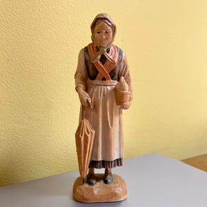 May include: A carved wooden figurine of a woman in traditional attire, holding a closed umbrella and a container. The figure is painted in shades of brown, beige, and red, with intricate details in the clothing and facial features. The figurine stands on a small, carved base.