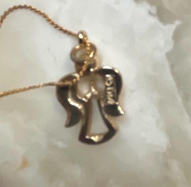 Pode incluir: Um colar de ouro com um pingente de anjo e uma corrente delicada. O pingente &eacute; detalhado com um design de asas de anjo e a palavra "WINGS" est&aacute; gravada na parte de tr&aacute;s.