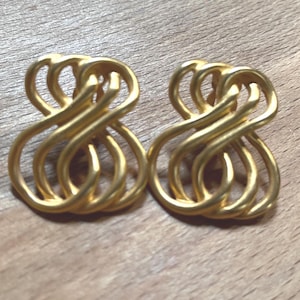 May include: A pair of gold-toned earrings featuring an openwork design of interlocking, fluid loops. The metal has a matte finish and a symmetrical, abstract shape that creates a continuous, woven pattern across the surface of each earring.