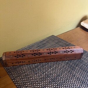 May include: A long, hexagonal wooden incense burner box featuring intricate laser-cut patterns along its sides. The brown wood surface is decorated with small, inlaid star-shaped metal accents, creating a detailed, geometric design throughout the piece.