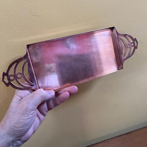 May include: A rectangular copper tray with decorative handles. The tray has a polished surface and a simple, elegant design. The handles feature an openwork design. The tray is held by a person.