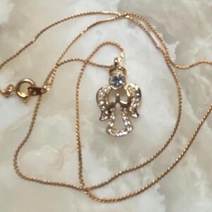 Pode incluir: Um colar de corrente de ouro com um pequeno pingente de anjo. O anjo &eacute; adornado com strass transparentes e tem um &uacute;nico strass maior no centro.