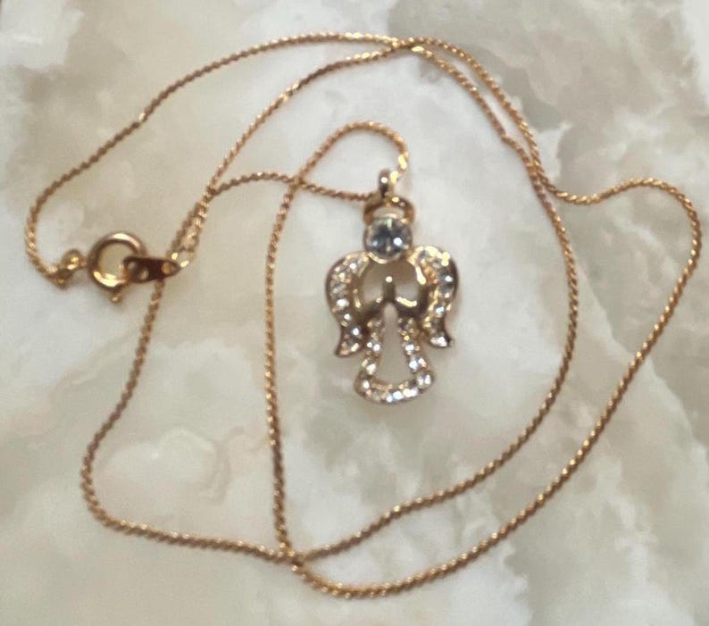Pode incluir: Um colar de corrente de ouro com um pequeno pingente de anjo. O anjo &eacute; adornado com strass transparentes e tem um &uacute;nico strass maior no centro.