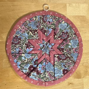 May include: A round, pink and white quilted hot pad with a floral pattern and a star design in the center. The hot pad has a loop for hanging.