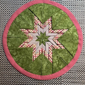 Könnte beinhalten: Ein runder, handgefertigter, gesteppter Platzdeckchen mit einem Sternmuster. Der zentrale Stern besteht aus grauem und gemustertem Stoff, umgeben von grünen Stoffsegmenten. Ein rosa Rand umrahmt das Design.