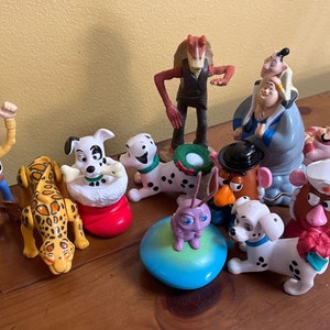 Può includere: Una collezione di 11 personaggi giocattolo di plastica, tra cui personaggi di Toy Story, 101 Dalmatiani, Star Wars e Mr. Potato Head. Le figure sono disposte su una superficie di legno.