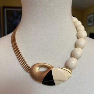 May include: A necklace featuring a multi-strand gold-tone snake chain on one side and a series of smooth cream-coloured spherical beads on the other. The two sides connect at a central focal piece with a brushed gold, black, and cream finish.