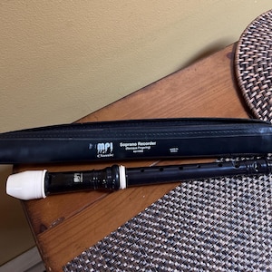 May include: A black soprano recorder with a white mouthpiece and a black case. The case is labeled "MPI Classic Soprano Recorder" with the Baroque fingering system. The recorder is lying on a wooden surface.