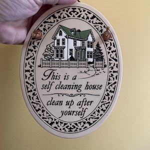May include: Oval ceramic plaque featuring a house illustration and the text "This is a self cleaning house clean up after yourself". The house has a green roof and white siding, with a decorative black border.