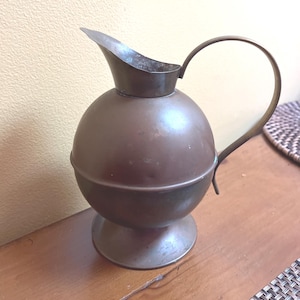 May include: A vintage copper-colored pitcher with a spherical body and a spout. It has a curved handle and sits on a small, round base. The pitcher is made of metal and has a tarnished appearance, suggesting age.