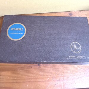 May include: A vintage Scrabble game box in dark gray with a blue and gold circular logo. The logo reads "SCRABBLE" and "A Crossword Game". The box also features the Sel-Right logo and the manufacturer's name.