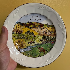 May include: A white decorative plate with a gold rim and a detailed countryside scene. The plate features a colorful illustration of people riding horses, trees, and a small village. The plate has a wheat stalk design around the rim.