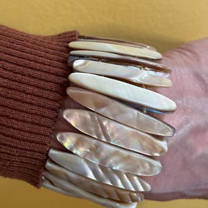 May include: A pearlescent bracelet with elongated, oval-shaped segments. The segments are in shades of cream, beige, and brown, with iridescent highlights. The bracelet is worn on a wrist, and the background is a solid yellow.