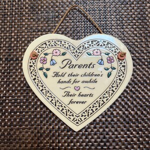 May include: A heart-shaped ceramic wall plaque in cream with a brown leather hanging cord. It features a decorative black filigree border, pink and blue floral accents, and the text: Parents Hold their children's hands for awhile Their hearts forever.