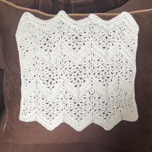 Pode incluir: Um cobertor branco de crochê com um padrão em zigue-zague. O cobertor é retangular com uma borda ondulada. O trabalho de crochê é detalhado, com espaços abertos criando uma textura delicada. O cobertor é exibido em uma almofada marrom.