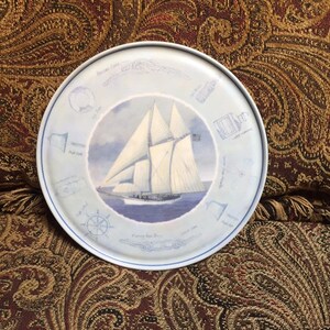 May include: A blue and white ceramic plate with a nautical theme. The plate features a sailboat with white sails and a blue hull. The plate is decorated with nautical symbols, including a compass, a ship's wheel, and a rope.