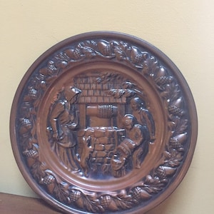 May include: A copper-colored decorative plate with a raised relief scene of figures near a well. The plate's border features a repeating pattern of acorns and leaves. The figures are dressed in period clothing.