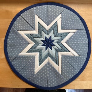 Könnte beinhalten: Ein runder, gesteppter Topfuntersetzer mit einem zentralen Sternmuster. Der Stern besteht aus verschiedenen Blautönen und weißen Akzenten. Der äußere Rand ist marineblau. Der Untersetzer hat einen Durchmesser von etwa 25 cm.