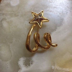 May include: A gold-toned brooch featuring a stylized star with a silver center and a swirling, ribbon-like design.