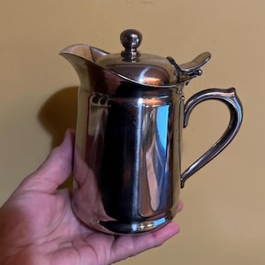May include: A silver-colored creamer with a lid and handle. The creamer has a spout for pouring and a small knob on top of the lid. The handle is curved, and the creamer is held in a hand.