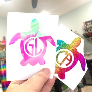 May include: Two turtle-shaped stickers. One is pink and green with the letters "GL" inside. The other is rainbow-colored with the letters "A" and a symbol inside. Both stickers are on white backing.