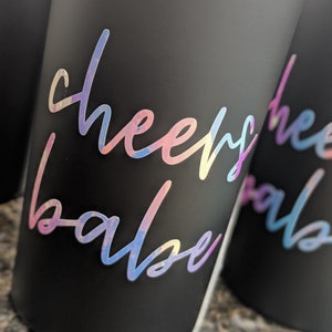 May include: Close-up of several black tumblers with iridescent "cheers babe" script. The text is in a cursive font and appears to be a sticker or decal. The tumblers are cylindrical and have a matte finish.