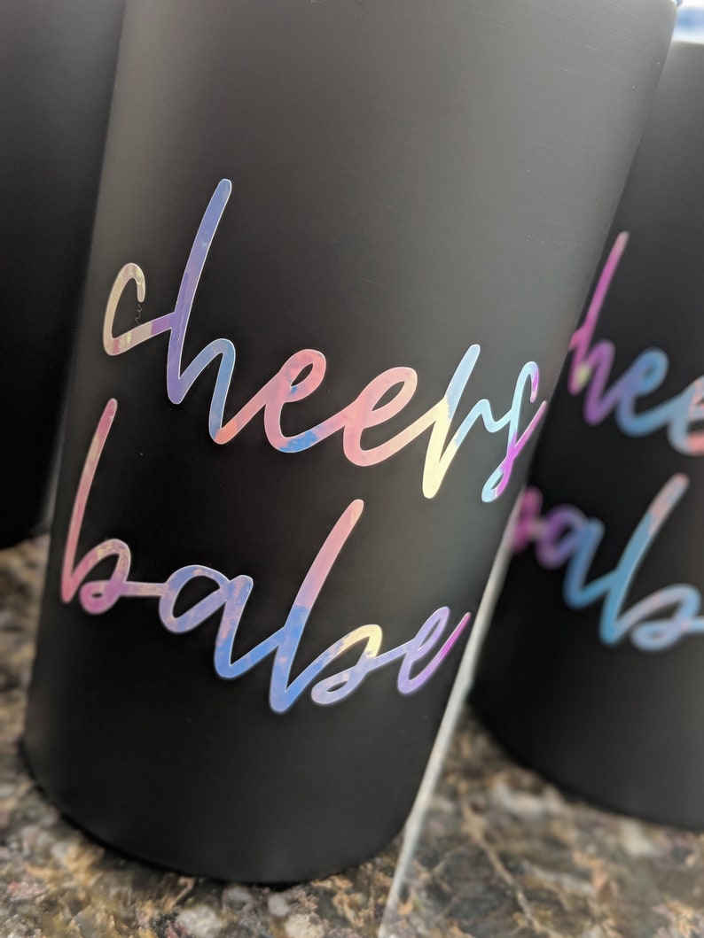 May include: Close-up of several black tumblers with iridescent "cheers babe" script. The text is in a cursive font and appears to be a sticker or decal. The tumblers are cylindrical and have a matte finish.