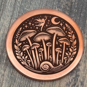 May include: A copper-colored round coin with a black engraved design. The design features mushrooms, a fairy, a crescent moon, stars, and a snail. The coin has a detailed, whimsical illustration.