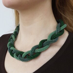 May include: A green and silver beaded necklace with a braided design. The necklace is made of small, round beads that are woven together in a complex pattern.