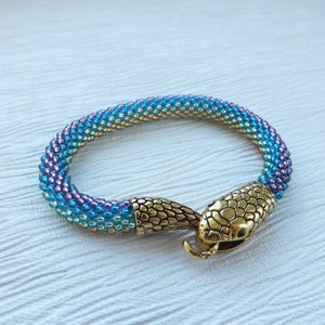 May include: A beaded bracelet with a gradient of blue, green, and gold colors. The bracelet has a snake-shaped clasp.
