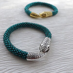 Puede incluir: Dos pulseras de cuentas con cierres de cabeza de serpiente. Una pulsera es verde azulado y la otra es dorada.