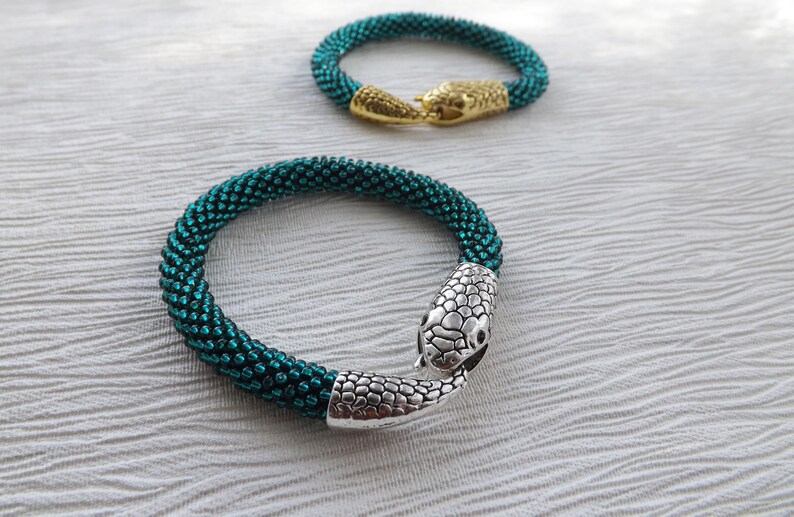 Puede incluir: Dos pulseras de cuentas con cierres de cabeza de serpiente. Una pulsera es verde azulado y la otra es dorada.