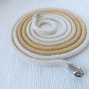 Puede incluir: Un collar largo de cuentas de m&uacute;ltiples hilos con un degradado de blanco a beige. El collar tiene un cierre en forma de serpiente plateado.