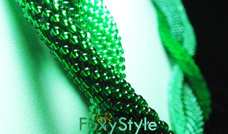May include: Close-up of a green beaded necklace with a gradient of darker green beads. The necklace is made of small, round beads that are tightly woven together.