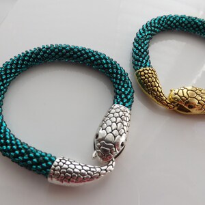 Puede incluir: Dos pulseras de serpiente de cuentas con cierres de cabeza de serpiente plateados y dorados. Las pulseras son de color verde azulado.