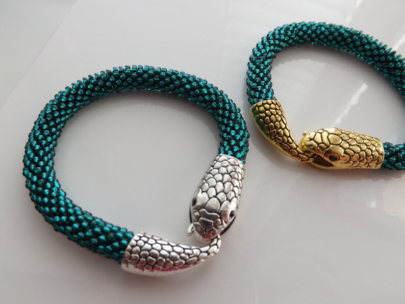 Puede incluir: Dos pulseras de serpiente de cuentas con cierres de cabeza de serpiente plateados y dorados. Las pulseras son de color verde azulado.