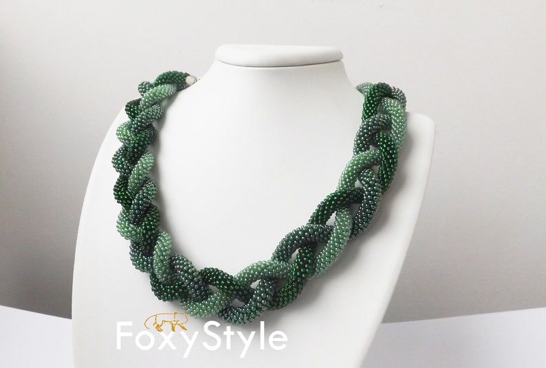 May include: A green beaded necklace with a gradient of shades from light to dark green. The necklace is made of small, round beads that are woven together in a braided pattern. The necklace is displayed on a white mannequin.