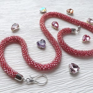 May include: A pink beaded necklace with a silver clasp and a silver chain. The necklace is made of small, round beads and is about 18 inches long. The necklace is surrounded by small, heart-shaped crystals.