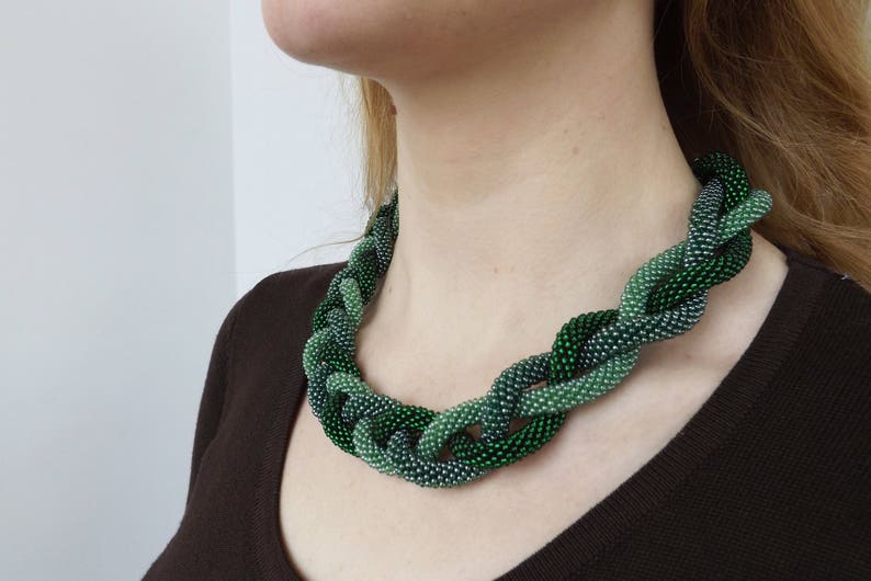May include: A green and silver beaded necklace with a braided design. The necklace is made of small, round beads that are woven together in a complex pattern.