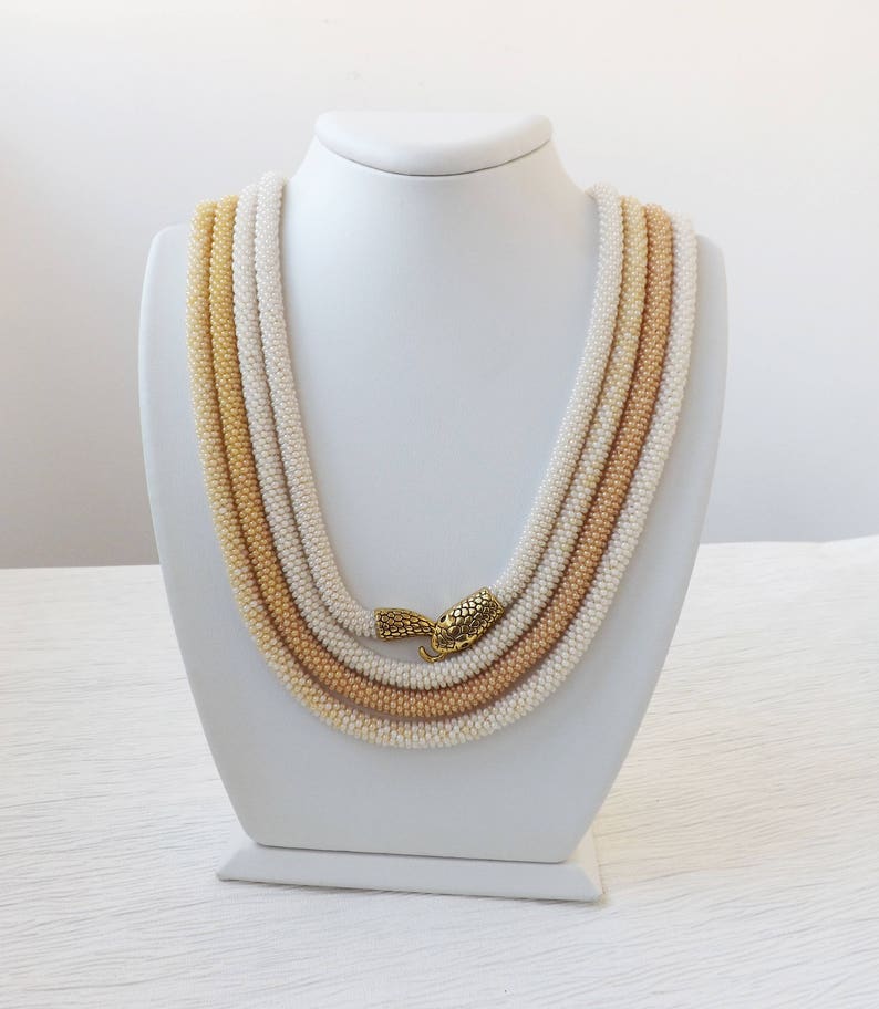 Puede incluir: Un collar de cuentas de m&uacute;ltiples hilos con un degradado de cuentas crema, beige y marr&oacute;n claro. El collar se muestra en un busto de maniqu&iacute; blanco y presenta un cierre dorado.