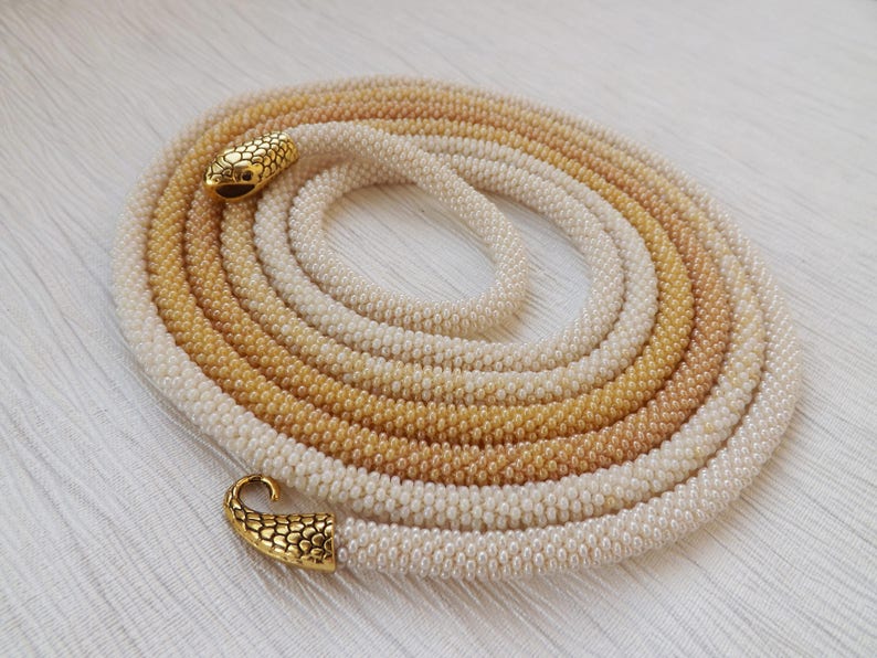Puede incluir: Un collar largo de cuentas con un degradado de colores que va del blanco al beige. El collar tiene dos cierres de serpiente dorados.