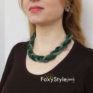 May include: A woman wearing a dark brown top with a green and blue beaded necklace. The necklace is a braided design with a subtle ombre effect. The text "FoxyStyle jewellery" is visible at the bottom of the image.