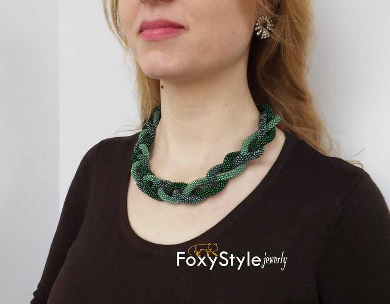 May include: A woman wearing a dark brown top with a green and blue beaded necklace. The necklace is a braided design with a subtle ombre effect. The text "FoxyStyle jewellery" is visible at the bottom of the image.