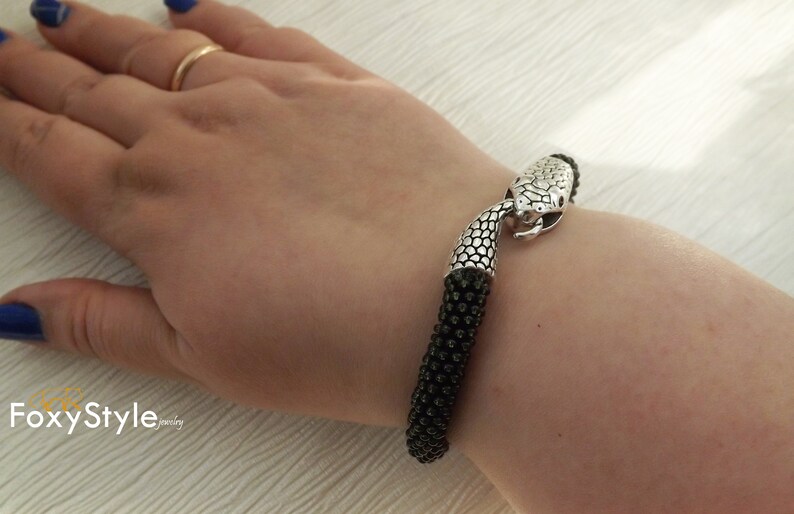 Puede incluir: Un brazalete negro de cuentas con un cierre de cabeza de serpiente plateado. El brazalete est&aacute; en la mu&ntilde;eca de una persona.