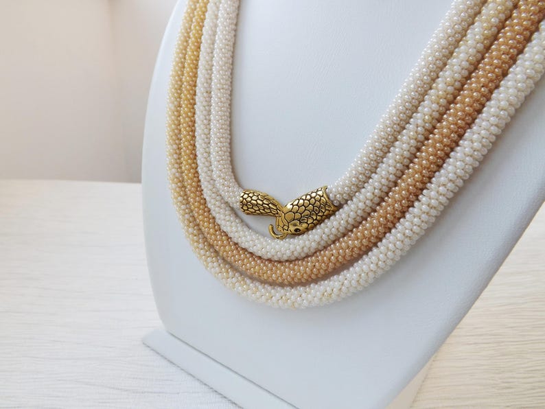 Puede incluir: Un collar de cuentas de m&uacute;ltiples hilos con un cierre dorado. El collar est&aacute; hecho de peque&ntilde;as cuentas blancas y crema.