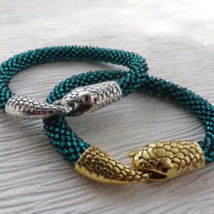 Puede incluir: Dos pulseras de cuentas con cierres de cabeza de serpiente. Una pulsera es verde azulado con un cierre plateado, la otra es verde azulado con un cierre dorado.