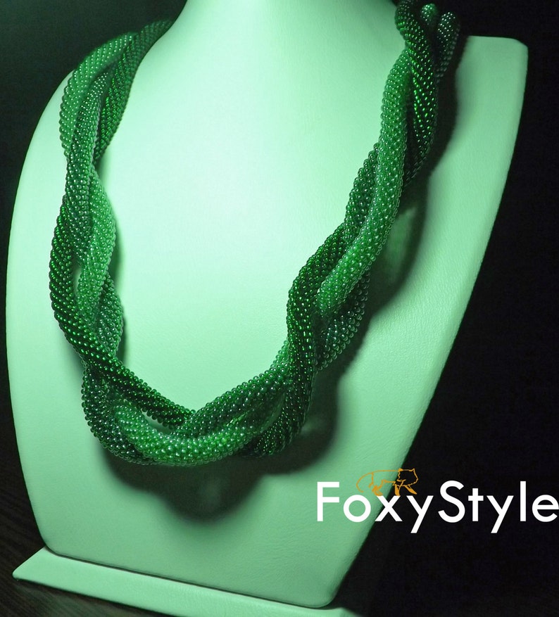 May include: A green beaded necklace with a twisted design. The necklace is displayed on a white mannequin bust.